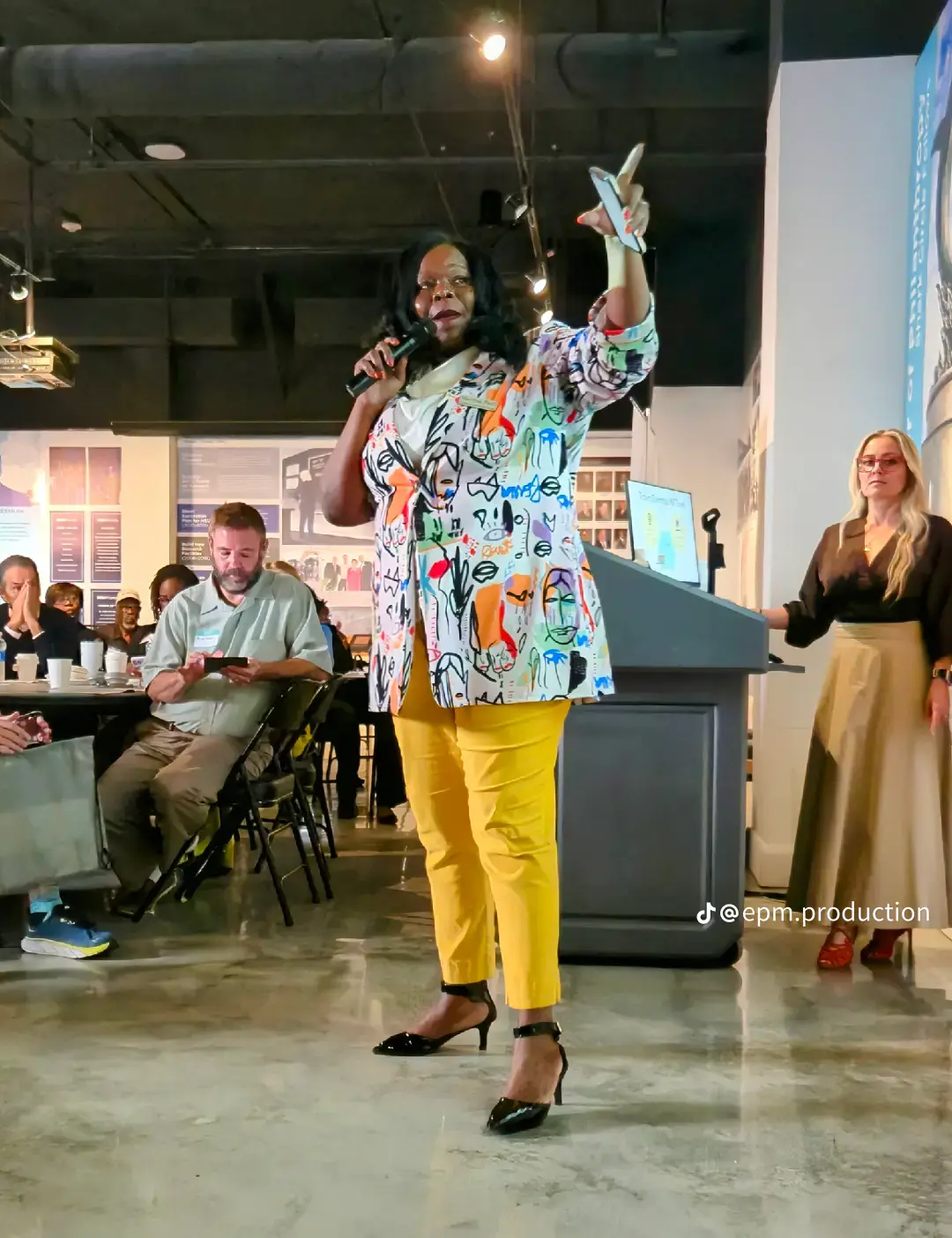 Marline Paul on stage in a colorful patterned blazer and yellow trousers, gesturing toward the audience