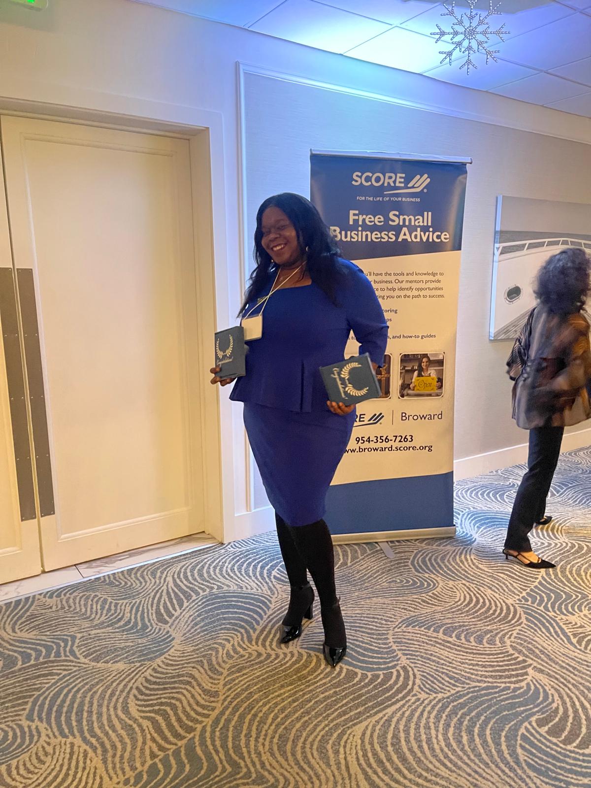 Marline Paul holding two awards beside the SCORE Broward banner