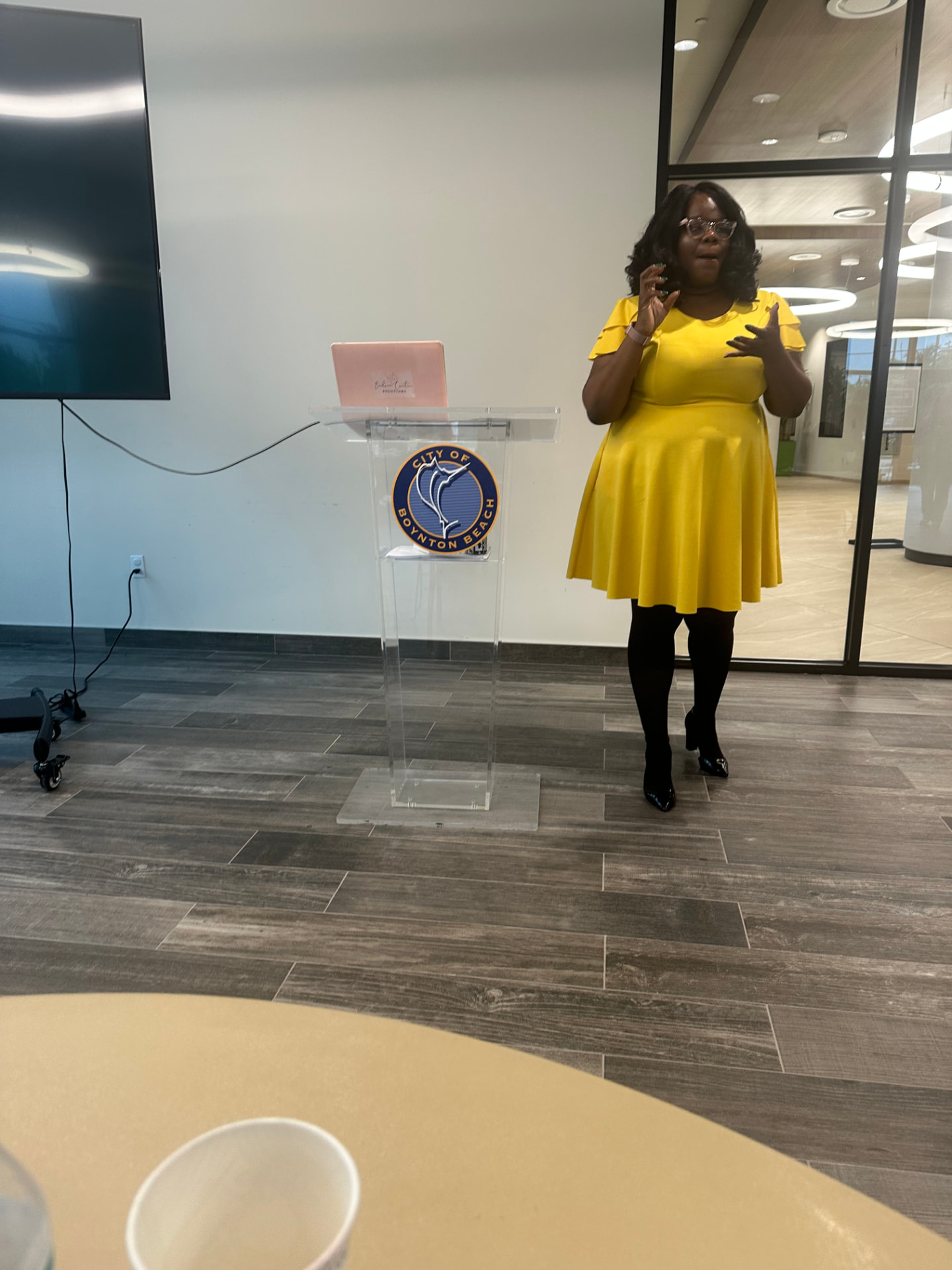 Marline Paul in a yellow dress presenting at the City of Boynton Beach Economic Development workshop beside a clear podium with the city seal